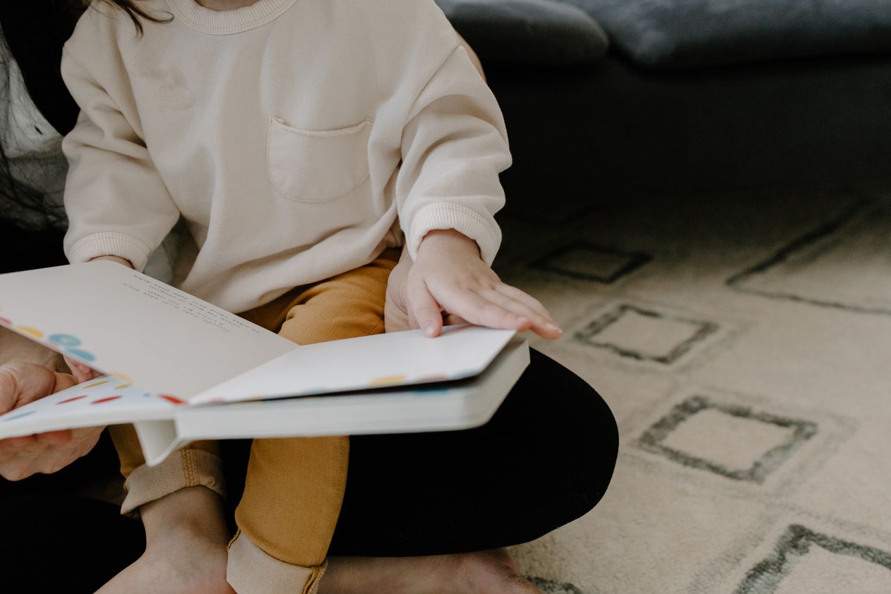 About A young child sitting on a carpet, reading a colorful picture book indoors.