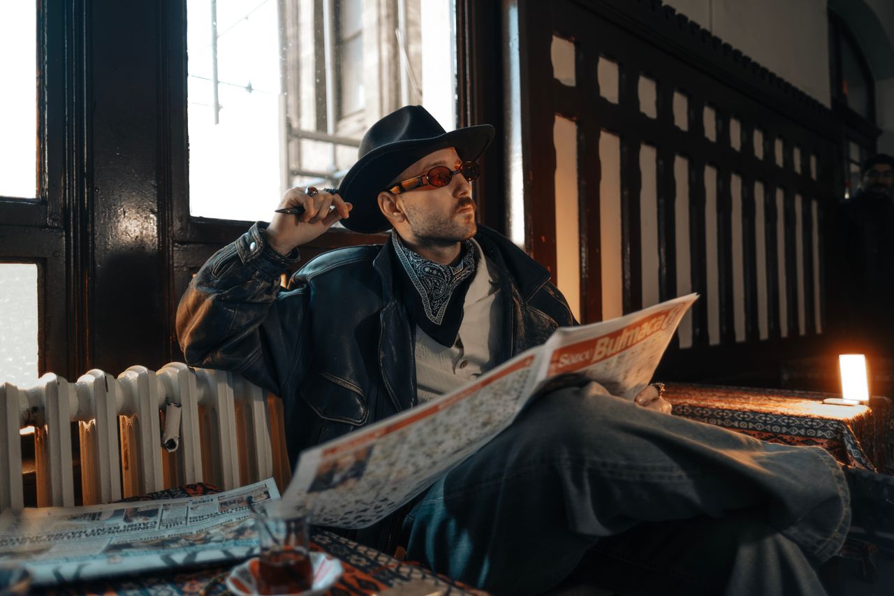 Stylish man in leather and hat reads newspaper in a vintage interior setting.